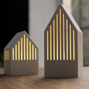 Op de afbeelding: Twee witte keramische kandelaars in de vorm van een huis. De huizen hebben een zadeldak en verticale uitgesneden lijnen die een warm, geel licht uitstralen. De kandelaars zijn verschillend van grootte, met de grotere aan de rechterkant.