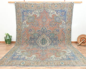 May include: A large, rectangular, vintage rug with an intricate floral pattern in shades of peach, blue, and green. The rug is displayed against a white wall and wooden floor, showcasing its detailed design and aged appearance.