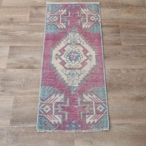 May include: A rectangular, vintage-style rug with a central diamond pattern in cream, surrounded by a field of faded rose and teal geometric designs. The rug is placed on a wooden floor.