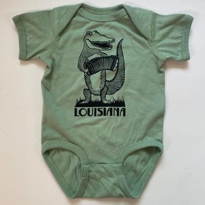 May include: A green baby onesie with a black and white illustration of an alligator playing an accordion. The onesie has the word "LOUISIANA" printed below the alligator.