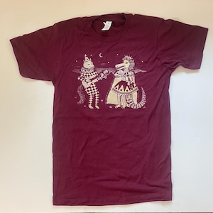 May include: Burgundy t-shirt featuring a whimsical illustration. The design depicts a cat playing a guitar and a crocodile wearing a crown and dress. The cat is dressed in a harlequin outfit. A crescent moon is in the sky.