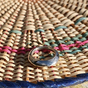 May include: A silver ring with a rounded, smooth surface. The ring is sitting on a woven mat with blue, pink, and green accents.