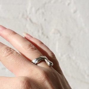 K&ouml;nnte beinhalten: Ein silberner Ring mit einem abstrakten, organischen Design. Der Ring wird an einem Finger getragen.
