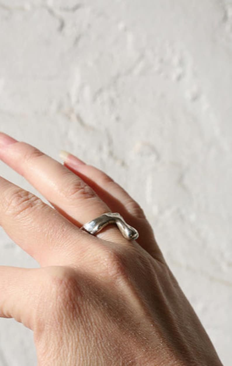 K&ouml;nnte beinhalten: Ein silberner Ring mit einem abstrakten, organischen Design. Der Ring wird an einem Finger getragen.