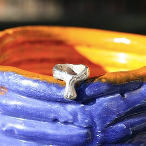 K&ouml;nnte beinhalten: Ein silberner Ring mit einem einzigartigen Design, das an einen Tropfen oder eine Tr&auml;ne erinnert. Der Ring liegt auf einer blauen und orangefarbenen Keramikfl&auml;che.