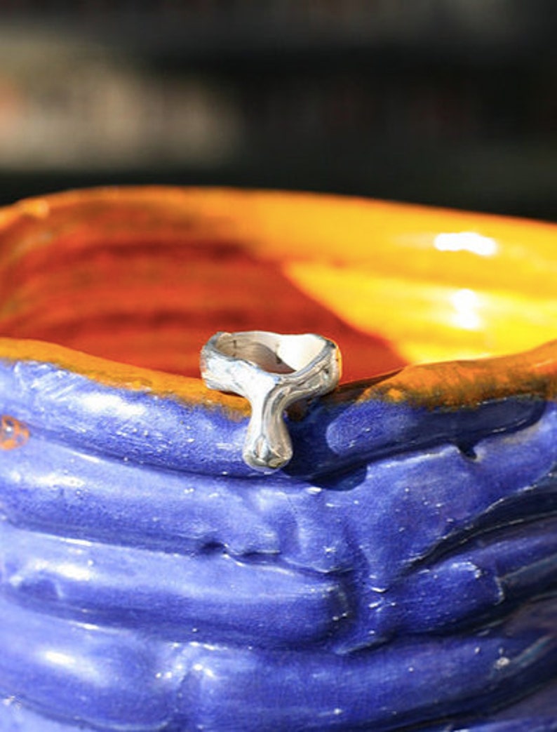 K&ouml;nnte beinhalten: Ein silberner Ring mit einem einzigartigen Design, das an einen Tropfen oder eine Tr&auml;ne erinnert. Der Ring liegt auf einer blauen und orangefarbenen Keramikfl&auml;che.