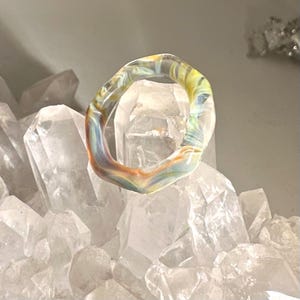 Peut inclure: Une bague claire et abstraite avec des tourbillons colorés de jaune, bleu et orange. La bague est posée sur un fond de cristaux de quartz clairs, créant un contraste saisissant. Le design de la bague est organique et fluide.