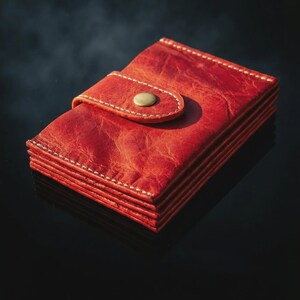 May include: A red leather wallet with a snap closure. The wallet is rectangular and has a stack of card slots. The stitching is white, and the snap is a brass color. The wallet is approximately 4 inches wide and 3 inches tall.