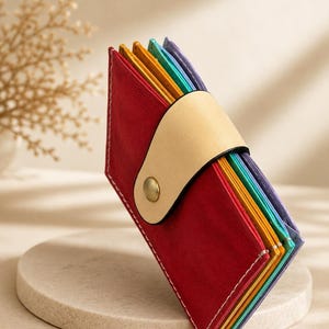 May include: A multi-colored leather accordion-style card holder featuring a red exterior panel, a beige leather strap closure with a brass snap button, and interior dividers in shades of yellow, teal, and purple with white stitching.