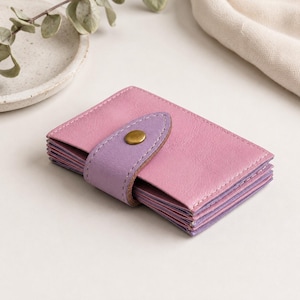 May include: A small, rectangular accordion-style card holder featuring a pink leather exterior and a lavender leather strap closure. The strap is secured with a single brass-toned metal snap button, and the interior compartments are visible in lavender.