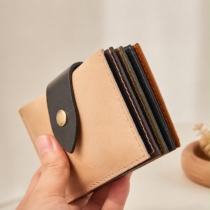 May include: A multi-compartment wallet featuring a light tan exterior panel secured by a black leather strap with a gold-toned metal snap button. The interior reveals several stacked card slots in varying shades of dark brown, green, and black.