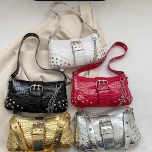 Puede incluir: Cinco bolsos de hombro de cuero sintético en negro, blanco, rojo, dorado y plateado. Cada bolso presenta una textura de cocodrilo, tachuelas plateadas, un detalle de cadena y un cierre de hebilla. Los bolsos tienen forma rectangular y correas ajustables.