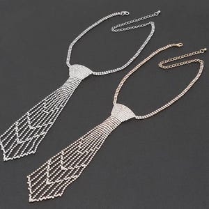 Peut inclure: Deux colliers en strass en forme de cravate. L'un est argenté, l'autre doré. Les deux ont une chaîne réglable. La partie cravate des colliers est composée de nombreux petits strass.