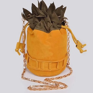 May include: A yellow suede pineapple-shaped bucket bag with a gold chain strap. The bag has a green suede top that resembles a pineapple crown.