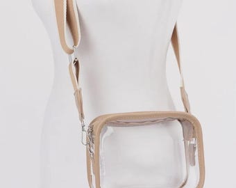 Puede incluir: Un bolso cruzado rectangular transparente con ribete y correa beige. El bolso tiene cierre de cremallera y herrajes plateados. La correa es una combinación de beige y blanco roto.