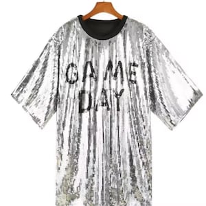 May include: Silver sequin t-shirt with the words "GAME DAY" in black block letters. The shirt has a black crew neck and short sleeves. The shirt is hanging on a wooden hanger.