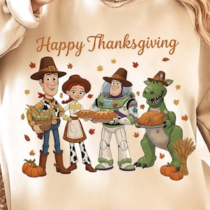 Puede incluir: Sudadera beige con el texto "Happy Thanksgiving" y un gráfico de personajes animados. Los personajes visten atuendos de Acción de Gracias y sostienen comida. Hojas de otoño esparcidas alrededor de los personajes.