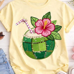 Puede incluir: Camiseta amarilla clara con un diseño bordado de una bebida de coco. El diseño incluye una flor de hibisco rosa, hojas verdes y una pajita a rayas. El coco tiene parches verdes. Ropa casual.