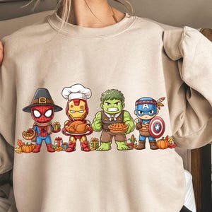 Peut inclure: Sweat-shirt beige avec un motif de Thanksgiving. Le graphisme représente Spider-Man, Iron Man, Hulk et Captain America, chacun tenant des objets festifs. Le motif comprend des citrouilles et des feuilles d'automne.