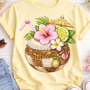 May include: A light yellow t-shirt featuring a detailed graphic of a tropical cocktail in a coconut shell. The design includes a pink hibiscus flower, lime, and a small pineapple. A pink straw and a tiny umbrella complete the summery look.