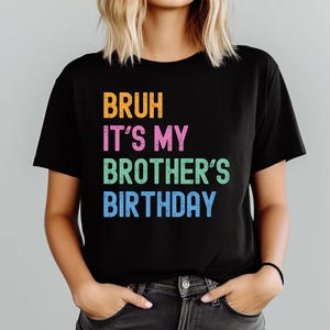 May include: Black crew neck t-shirt featuring the phrase "BRUH IT'S MY BROTHER'S BIRTHDAY" in orange, pink, green, and blue. The shirt is made of a soft material.