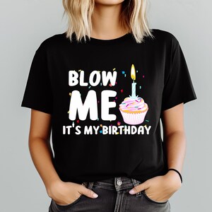 May include: Black t-shirt with the text "BLOW ME IT'S MY BIRTHDAY" in white letters. A cupcake with a lit candle is next to the text. Colorful confetti is scattered around the text and cupcake.