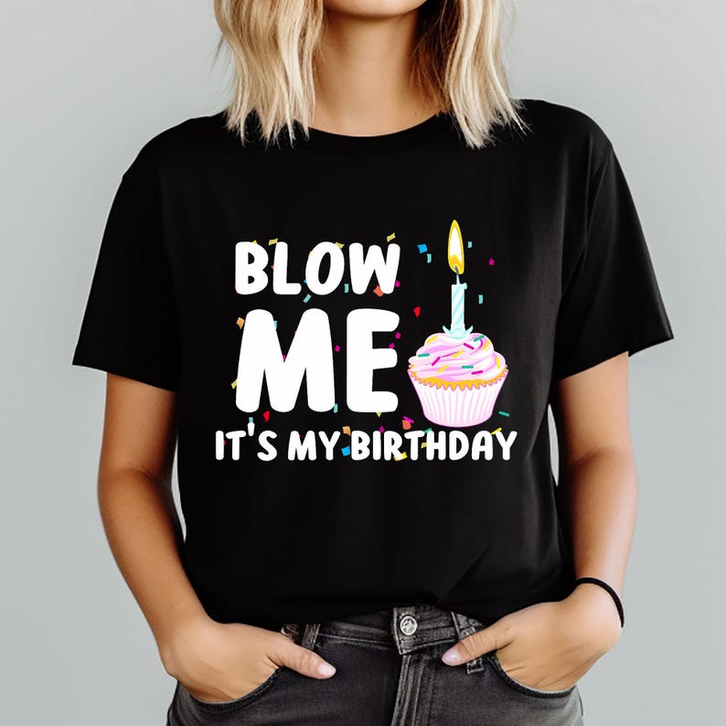 May include: Black t-shirt with the text "BLOW ME IT'S MY BIRTHDAY" in white letters. A cupcake with a lit candle is next to the text. Colorful confetti is scattered around the text and cupcake.