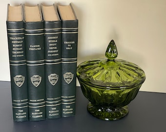 May include: Four dark green hardcover books from The Harvard Classics series, with gold lettering and the Harvard University crest. A green glass candy dish with a lid sits to the right.