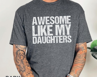 May include: Dark heather gray t-shirt with the text "AWESOME LIKE MY DAUGHTERS" in white block letters. The shirt is a crew neck style and has short sleeves. The words "DARK HEATHER" are printed at the bottom of the image.