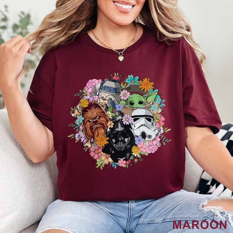 Puede incluir: Camiseta granate con un dise&ntilde;o de corona floral con personajes de Star Wars: Chewbacca, Darth Vader, un Stormtrooper, R2-D2 y Grogu. La palabra "MAROON" est&aacute; impresa en la esquina inferior derecha.