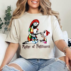 Puede incluir: Camiseta beige con una ilustraci&oacute;n colorida de un personaje con cabello rojo y un vestido de retazos, con dos ni&ntilde;os esqueleto. El texto "Mother of Nightmares" est&aacute; impreso debajo. La camiseta es de material suave.