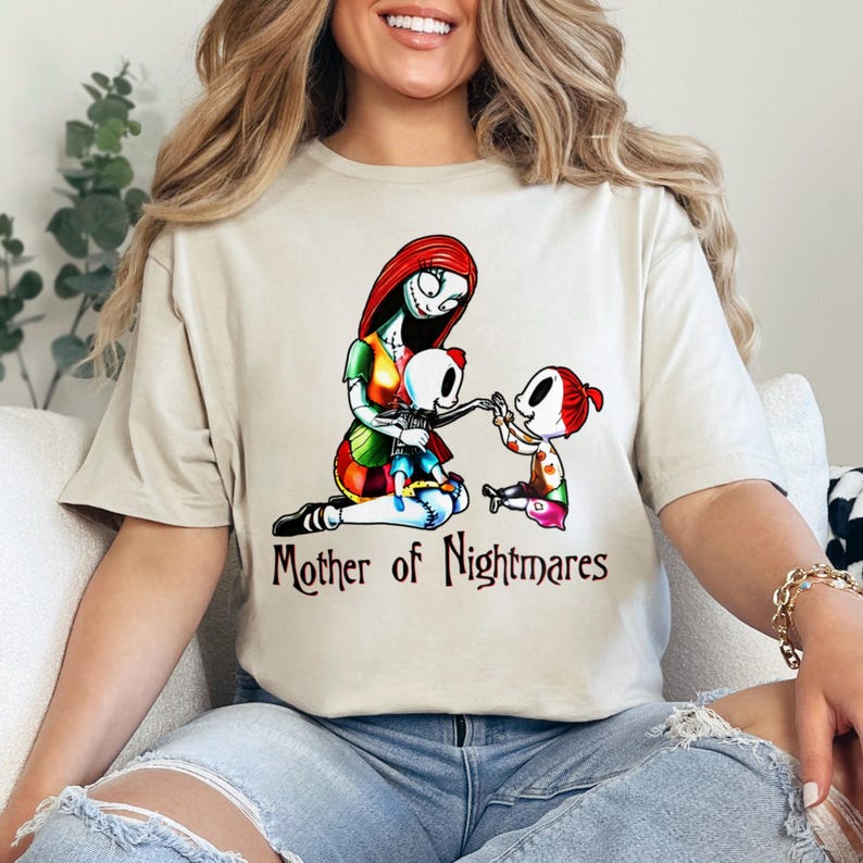 Puede incluir: Camiseta beige con una ilustraci&oacute;n colorida de un personaje con cabello rojo y un vestido de retazos, con dos ni&ntilde;os esqueleto. El texto "Mother of Nightmares" est&aacute; impreso debajo. La camiseta es de material suave.