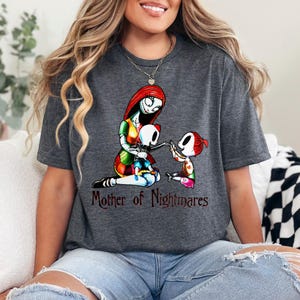 Puede incluir: Camiseta gris oscuro con una ilustraci&oacute;n colorida de un personaje con cabello rojo y un vestido de retazos, con dos figuras esquel&eacute;ticas. El texto "Mother of Nightmares" est&aacute; impreso debajo. Prenda de vestir informal.