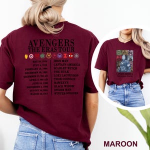 May include: Maroon t-shirt with "AVENGERS THE ERAS TOUR" text, character icons, and dates. The back lists characters such as Iron Man and Scarlet Witch. The front displays a Captain America graphic. The shirt is a deep red colour.