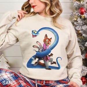 Op de afbeelding: Zandkleurige sweatshirt met een kleurrijke afbeelding van stripfiguren, waaronder een vos, een konijn en een blauwe slang. De afbeelding is gecentreerd op de voorkant van de top met lange mouwen. Het woord "SAND" staat linksonder in het wit gedrukt.