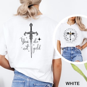May include: White t-shirt featuring a black sword design with the words "You do not yield." The shirt also has a round graphic with the text "BASGIATH WAR COLLEGE" and a dragon design. The word "WHITE" is at the bottom.