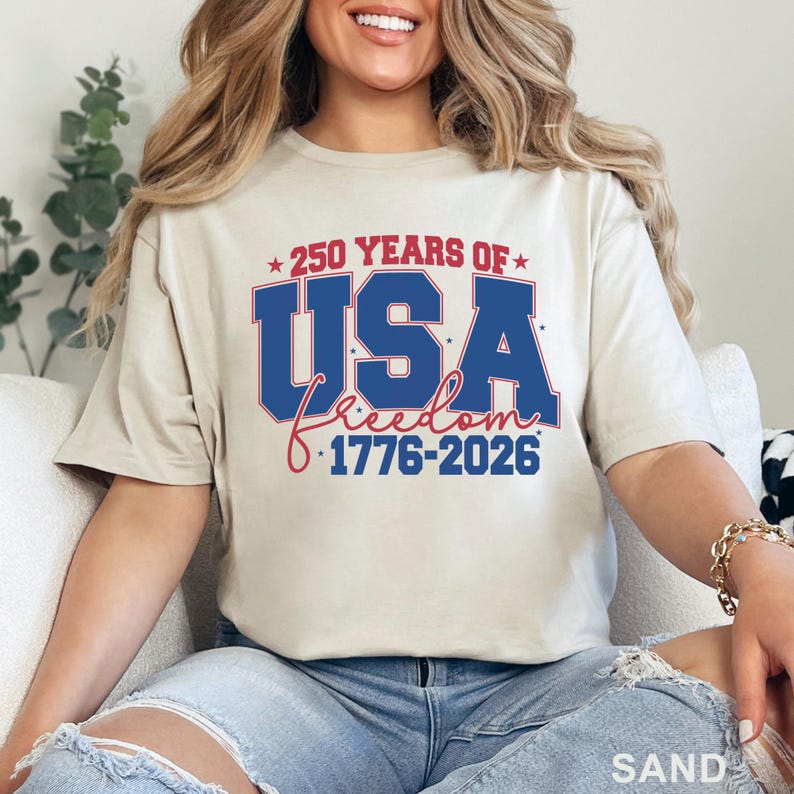 May include: Sand-colored t-shirt with a patriotic design. The graphic reads "250 YEARS OF USA freedom 1776-2026" in red and blue. The shirt is a crew neck and has short sleeves.