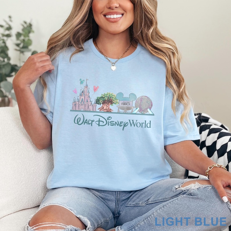May include: Light blue t-shirt featuring a Walt Disney World graphic. The design includes a castle, tree, and other park icons. The text "Walt Disney World" is printed below the graphic. The shirt is a light blue color.