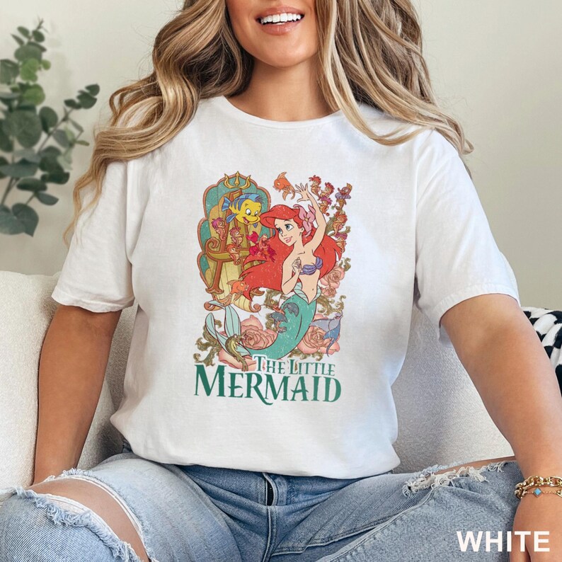 May include: White t-shirt featuring a colorful graphic of Ariel from The Little Mermaid, with the text "The Little Mermaid" in green. The design includes Ariel, Flounder, and floral elements. The word "WHITE" is printed at the bottom.