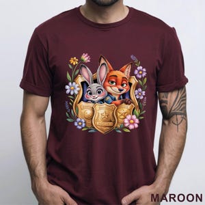 Puede incluir: Camiseta granate con una ilustraci&oacute;n de dibujos animados de Judy Hopps y Nick Wilde de Zootopia dentro de una placa de polic&iacute;a dorada, rodeada de flores de colores. La palabra "POLICE" es visible en la placa.
