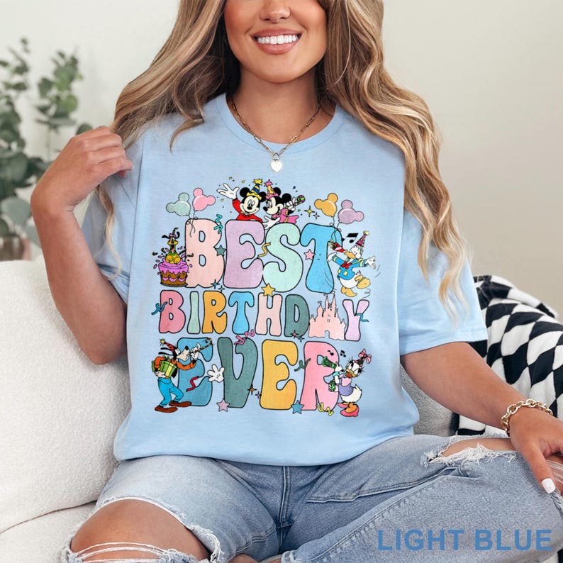 May include: Light blue t-shirt with the text "BEST BIRTHDAY EVER" in colorful bubble letters. The design includes cartoon characters like Mickey and Minnie Mouse, Donald and Daisy Duck, and Goofy. The shirt is a light blue color.