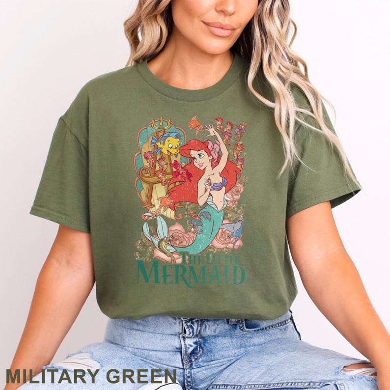 May include: Military green t-shirt featuring a colorful graphic of Ariel from The Little Mermaid. The design includes Ariel, Flounder, and floral elements. The text "THE LITTLE MERMAID" is printed below the graphic.