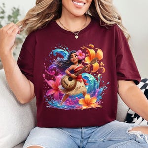 May include: Burgundy t-shirt featuring a vibrant illustration of a woman playing a ukulele, surrounded by colorful flowers and water splashes. The design includes a floral headband and a traditional skirt. The overall theme is tropical and musical.