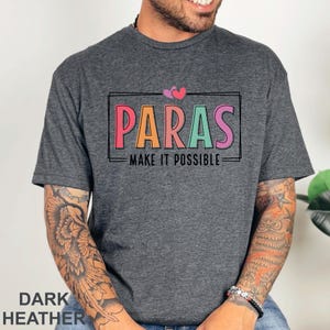 May include: Dark heather gray t-shirt with the word "PARAS" in colorful letters, framed by a black rectangle. The text "MAKE IT POSSIBLE" is below. Two pink hearts are above the word "PARAS".