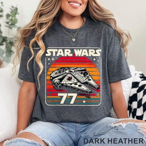 May include: Dark heather gray t-shirt featuring a Star Wars design. The graphic includes the text "STAR WARS" above a retro-style image of the Millennium Falcon spaceship set against a sunset. The number "77" is below the spaceship.