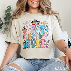May include: Sand-colored t-shirt with the phrase "BEST BIRTHDAY EVER" in colorful, retro-style lettering. The design includes cartoon characters like Mickey and Minnie Mouse, Donald Duck, and Goofy, with a birthday cake and castle.