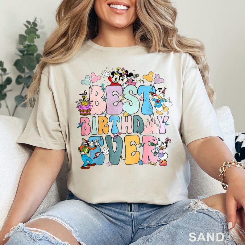 May include: Sand-colored t-shirt with the phrase "BEST BIRTHDAY EVER" in colorful, retro-style lettering. The design includes cartoon characters like Mickey and Minnie Mouse, Donald Duck, and Goofy, with a birthday cake and castle.