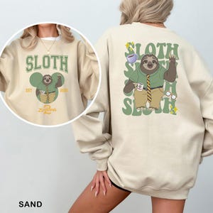 May include: Sand-colored sweatshirt featuring a cartoon sloth character from Zootopia. The front design includes the word "SLOTH" above the sloth graphic, with "EST 2015" and "Zootopia 2" below. The back has a larger sloth graphic with the word "SLOTH" repeated.