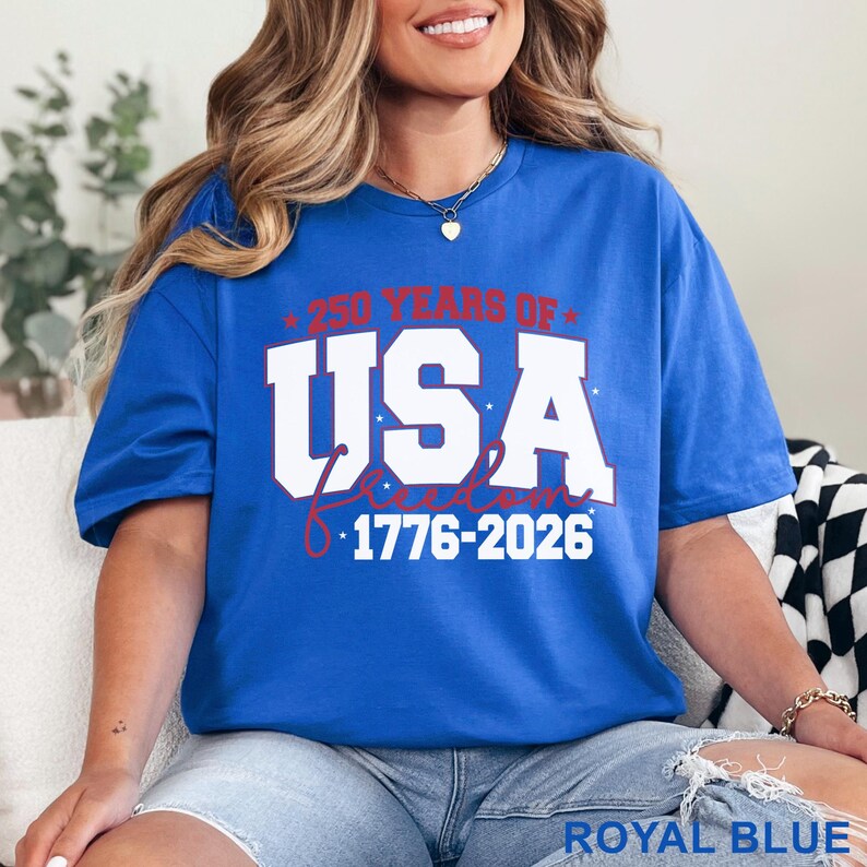 May include: Royal blue t-shirt with the text "250 YEARS OF USA FREEDOM 1776-2026" in white and red. The shirt is a solid color and has a relaxed fit. The text is centered on the front of the shirt.