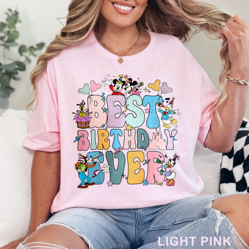 May include: Light pink t-shirt with the text "BEST BIRTHDAY EVER" in colorful, bubble letters. The design includes cartoon characters like Mickey Mouse, Donald Duck, and Goofy. The shirt is a crew neck style.